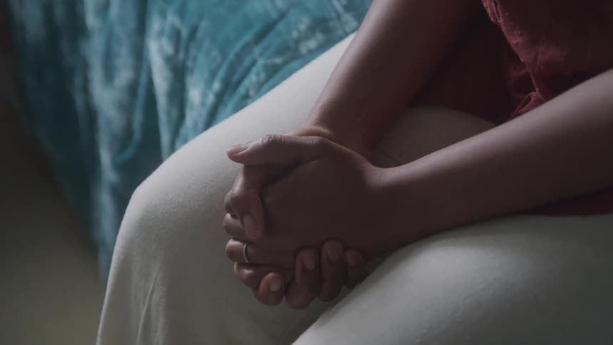The hands of a person closed and sitting on a bed and wearing white pants