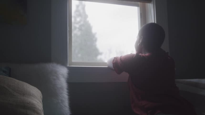 A woman wearing a red shirt with textured patterns leans on a windowsill gazing outside at lush green foliage through a window