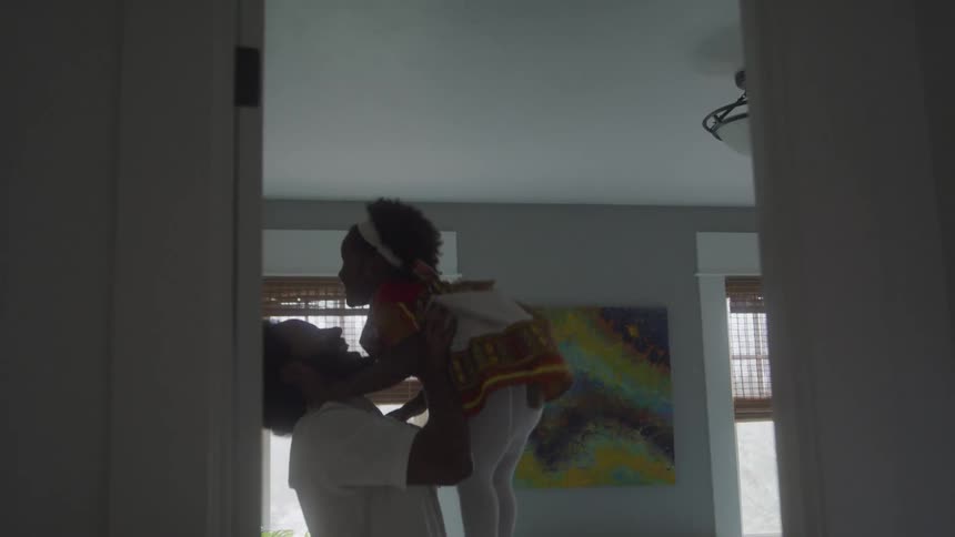 A person lifts a child dressed in a vibrant patterned outfit into the air inside a room with a window with blinds and a colorful abstract painting on the wall