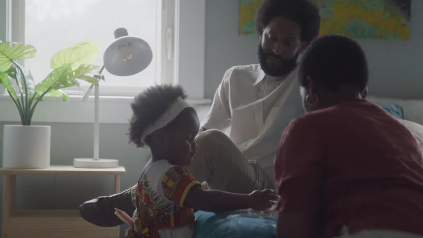 The father and mother sit together in a bright room while their daughter plays with toys on the bed