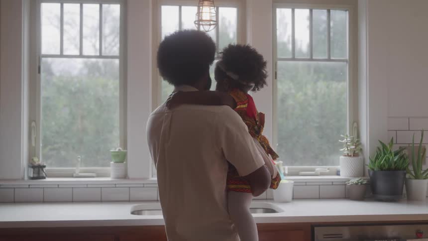A man standing in a kitchen holding his daughter on his lap and they are looking out the window