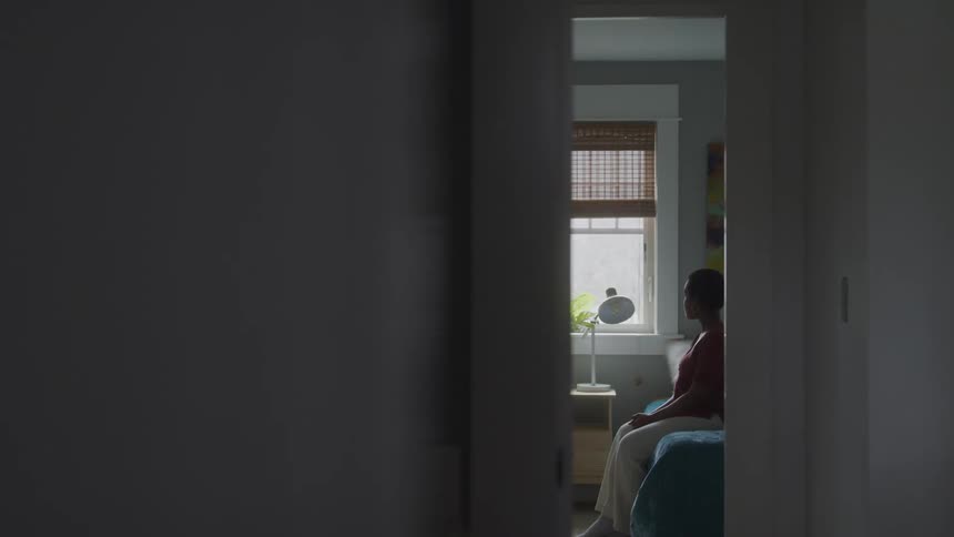 A woman in a red shirt is just sitting on a bed in a room and looking out the window