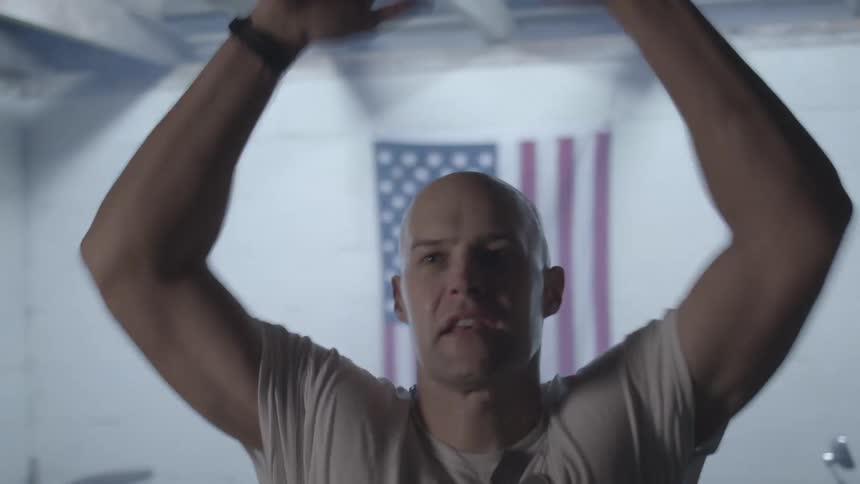 Bald man in tee shirt and dog tags does jumping jacks in front of a us flag