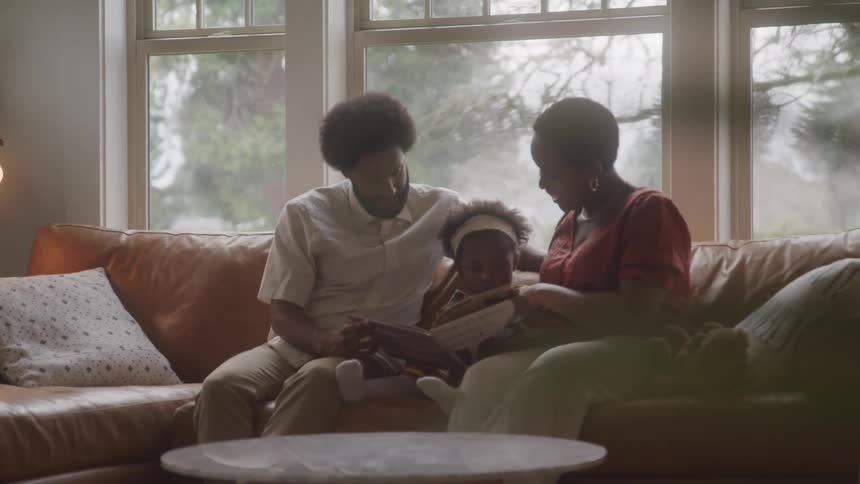 An adult man and woman are sitting on the couch reading a book to a little girl who is in between them