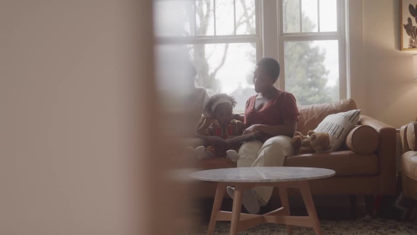 A family of three sits together on a brown couch in a living room with large windows and a round wooden coffee table in the foreground