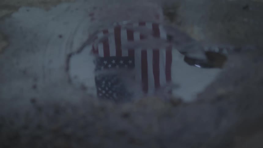 An us flag reflecting in a puddle of water and there are legs of a person moving in front