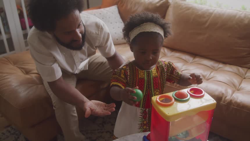 A child plays with a shapesorting toy while an adult assists both seated on a beige couch