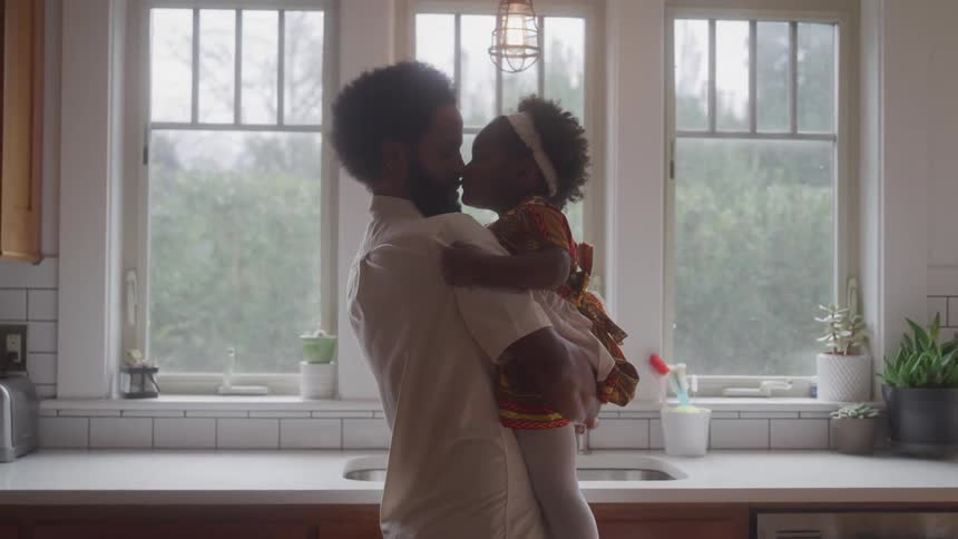 Father holds toddler daughter in a kitchen while she gives him a kiss on the nose