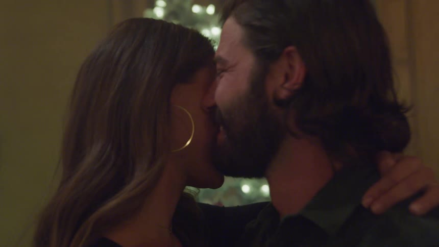 A young happy couple dances closely together in a room with a decorated christmas tree glowing in the background