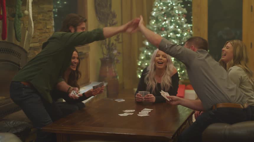 A group of friends sit around a wooden table playing cards with two of them exchanging a highfive and a decorated christmas tree glowing in the background