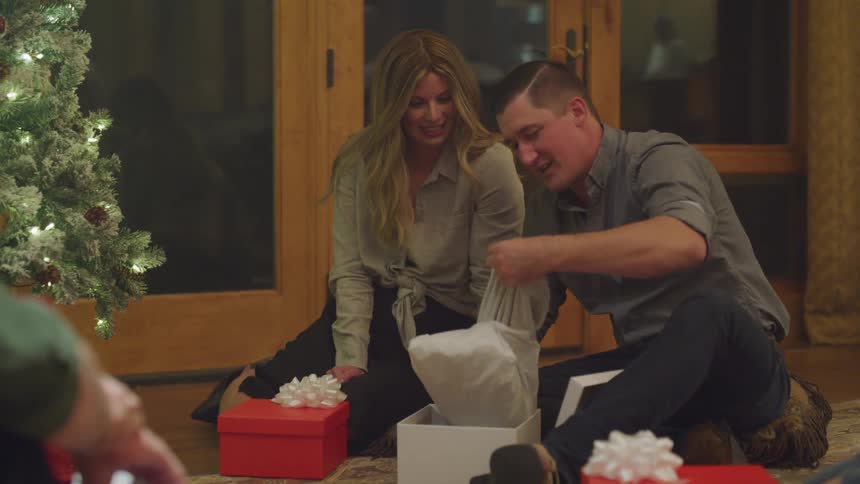 Adults sitting on the floor are opening presents near a christmas tree