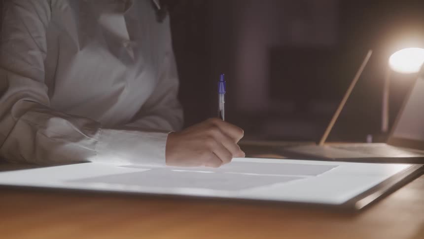 One person works at a light table drawing on a sheet of paper with a pen
