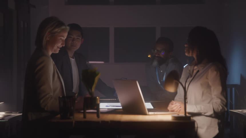 A group of people stands around a light table in a dimly lit room discussing while laptops and papers are spread across the table