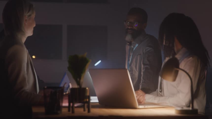A group of people stands around a light table in a dimly lit room discussing while laptops and papers are spread across the table