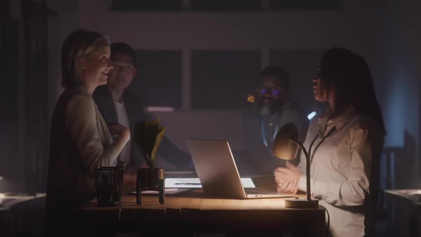A group of people stands around a light table in a dimly lit room discussing while laptops and papers are spread across the table
