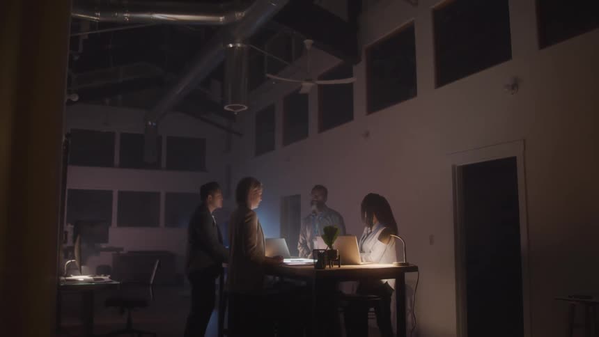 Four people are standing around a table in a dimly lit room with laptops and papers illuminated by the laptop screens soft glow