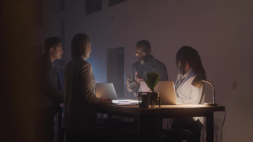 Four people standing around an office table with laptops open talking in a meeting