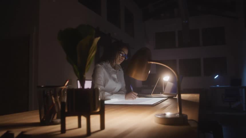 Woman writes at night while sitting at a desk in a dark room lit by a desk lamp