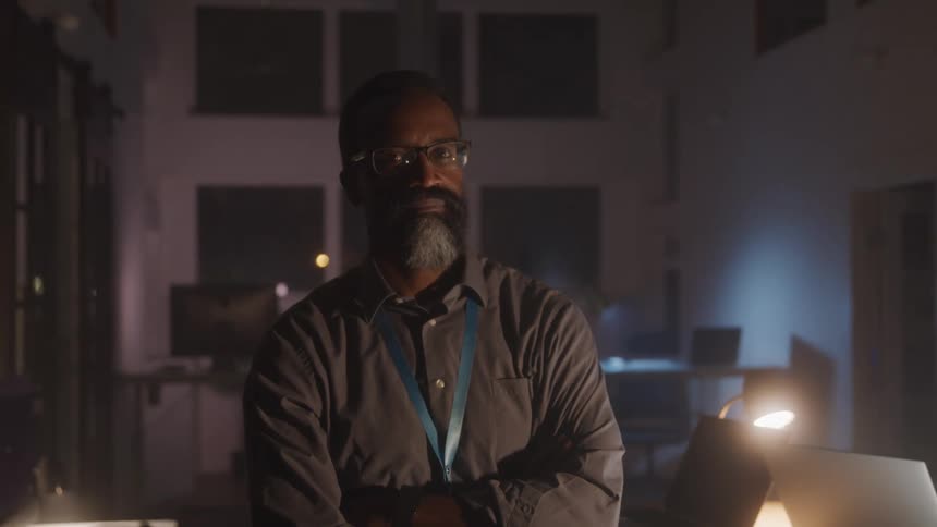 A man stands on a dimly lit office at night looking at the camera