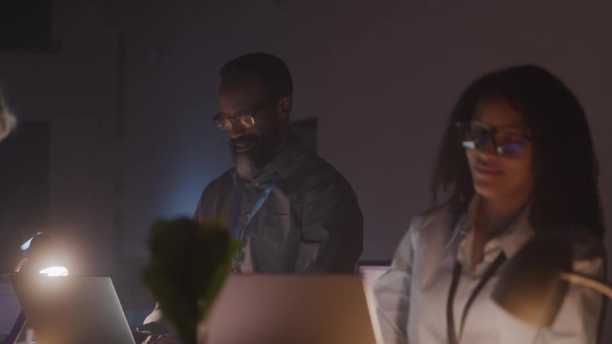Three individuals are working on laptops in a dimly lit room with a desk lamp providing focused illumination