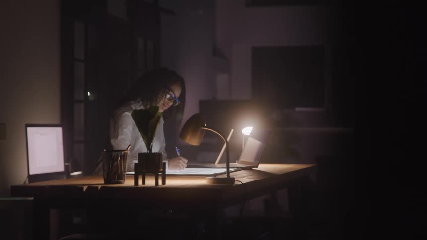 An illustrator works on a light table over a desk surrounded by lamps in a dimly lit office