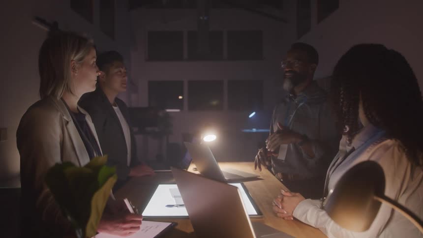 Group of four people in an office meeting in a dark room with laptop and tablets open on the table