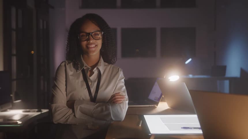 A woman stands beside a desk holding laptops and an illuminated drawing table in an office at night