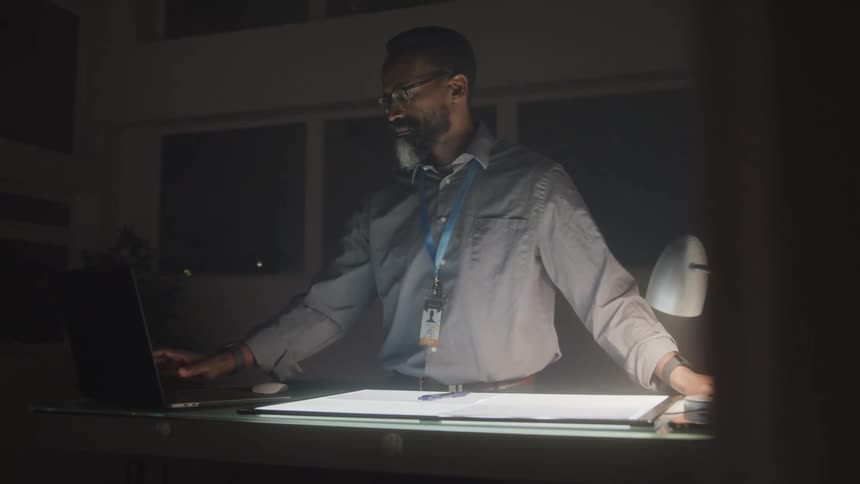 Man in an office room at night interacting with a lit screen on a desk next to a laptop
