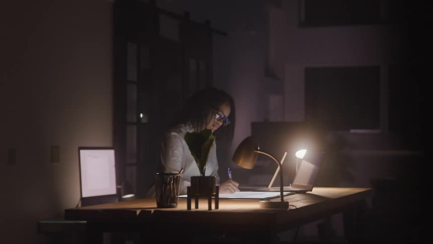 A woman works late at night in an office drawing on a light table with a laptop and desk lamp beside her