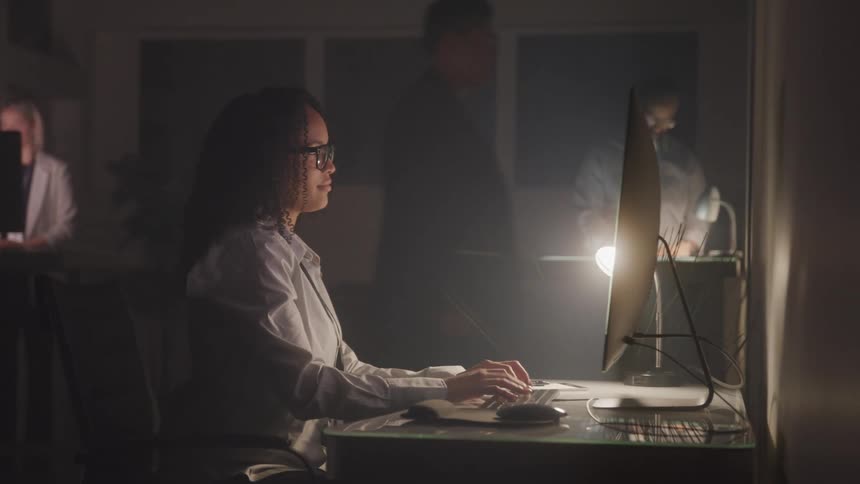 Woman sitting typing on a keyboard in an office room at night with other people