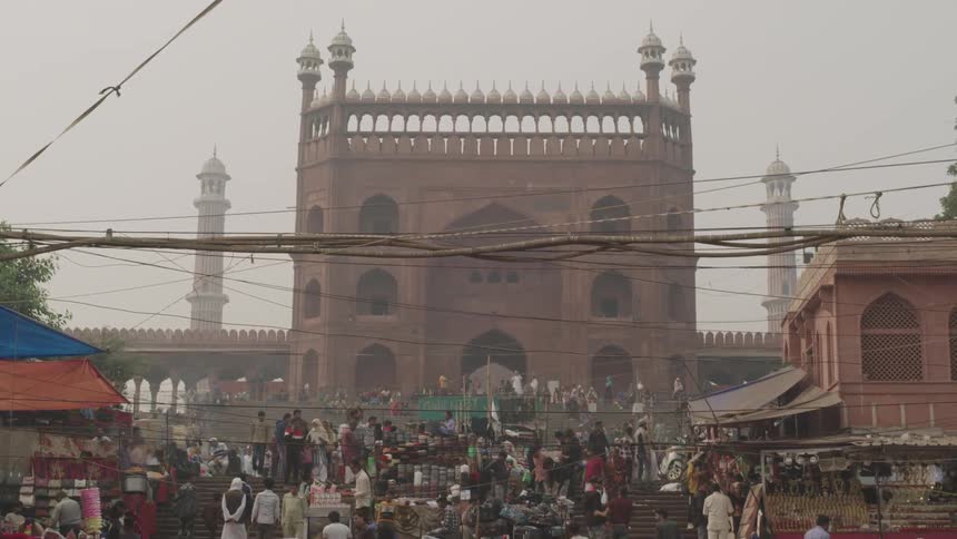 A large crowd of people is gathered on a series of steps likely at a market or event with an ornate mosque visible in the background