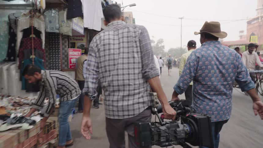 A film crew three men and a woman in casual clothing walking together down a crowded street lined with shops displaying fabric and textiles
