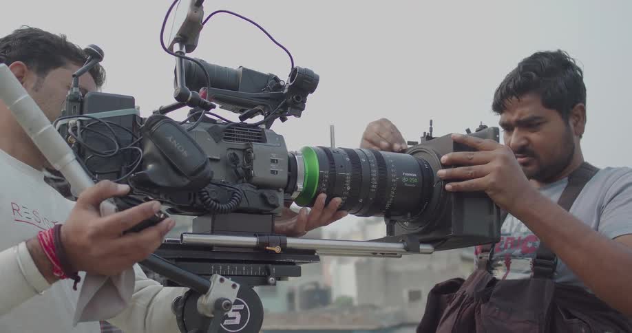 A film crew operating a professional camera on a stabilized rig while filming an outdoor scene with buildings visible in the background