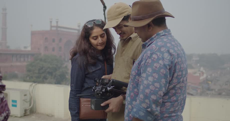 A filming crew gathers around the cameraman inspecting the camera with indian historic buildings in the background