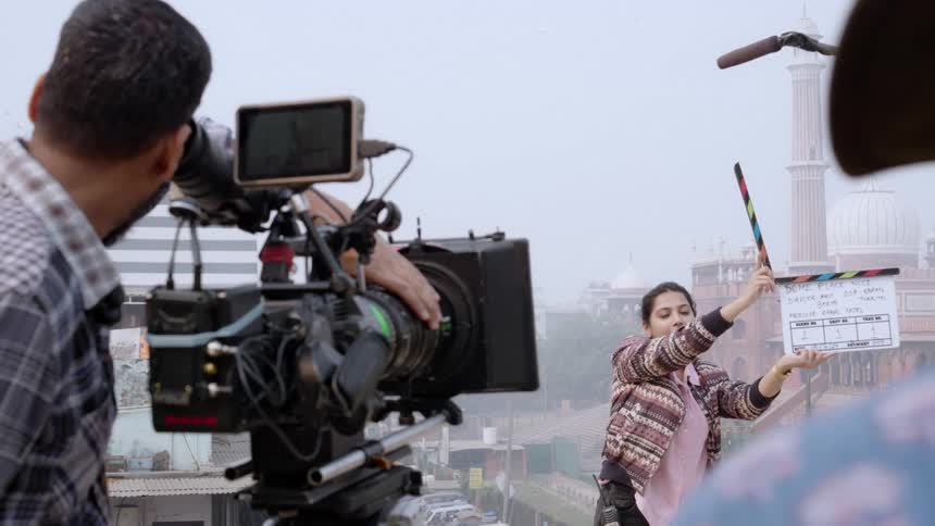 A film crew works on a rooftop operating cameras and equipment while one person holds a clapperboard in the background