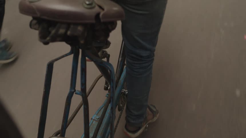 The rear of a vintage bicycle showing a brown leather seat blue frame and a persons legs pedaling