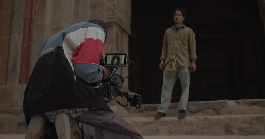 Camera operator kneels while taking video of a male model posing in front of a stone building
