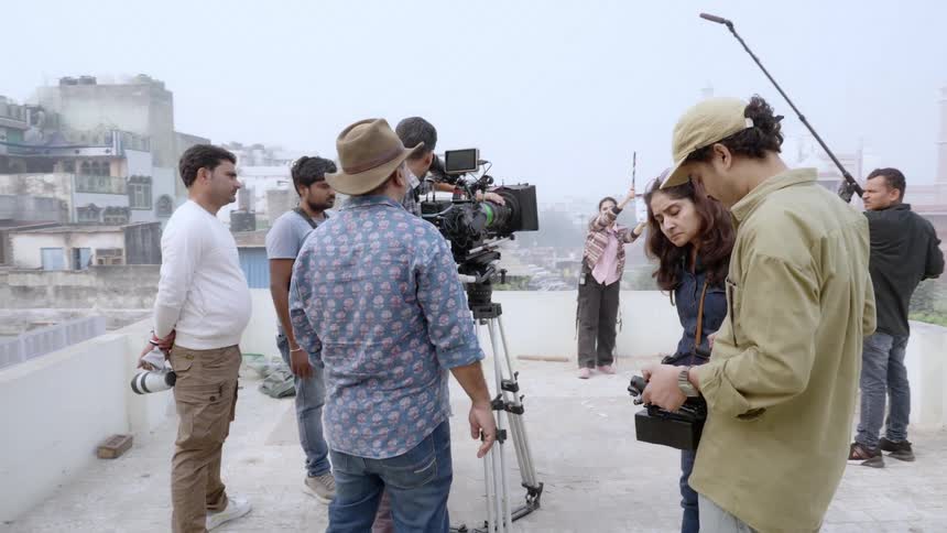 A film crew works on a rooftop operating cameras and equipment while one person holds a clapperboard in the background