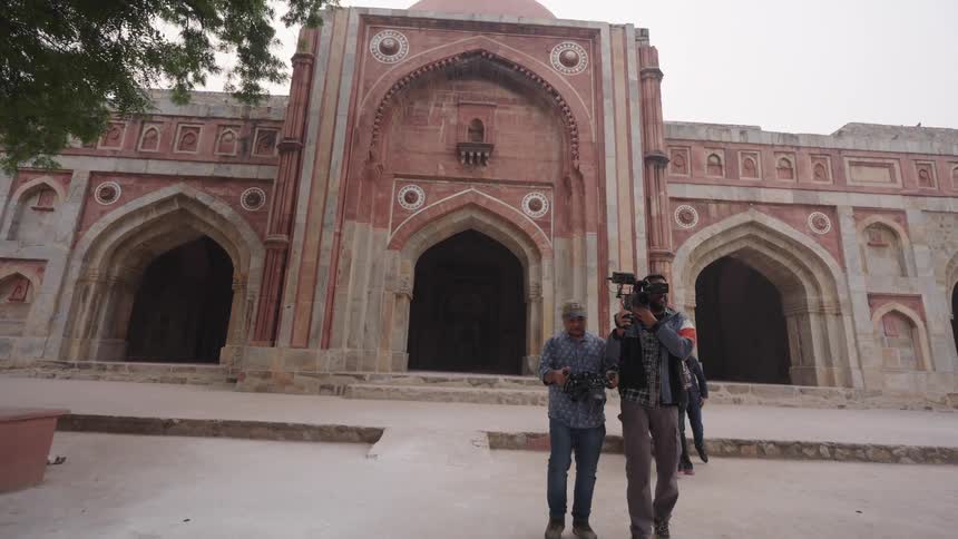 A film crew including a cameraman and a person holding a microphone stand in front of a historic building with arched doorways and a large central dome surrounded by trees