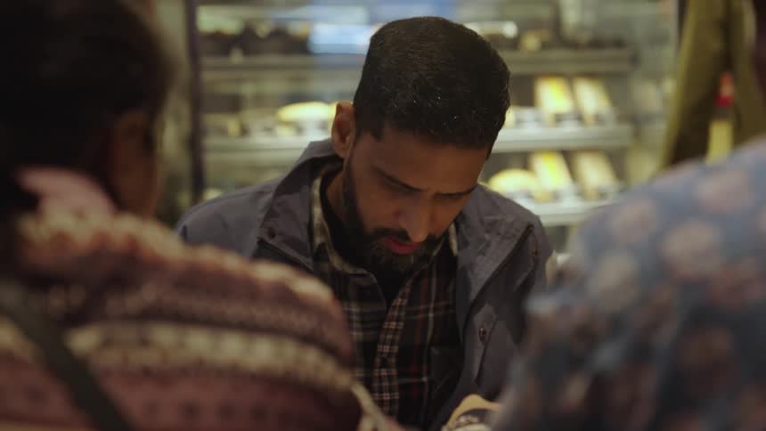 The man in the jacket is handed a pen by the woman sitting across from him with another man and the person in the green turns their back and walks away