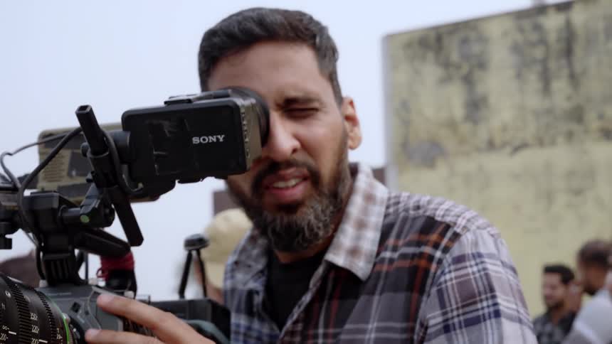 Male cameraman manipulates a video camera while a crowd stands behind him during the day