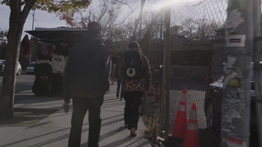 People walk along the sidewalk beside a wire fence on a bright sunny morning