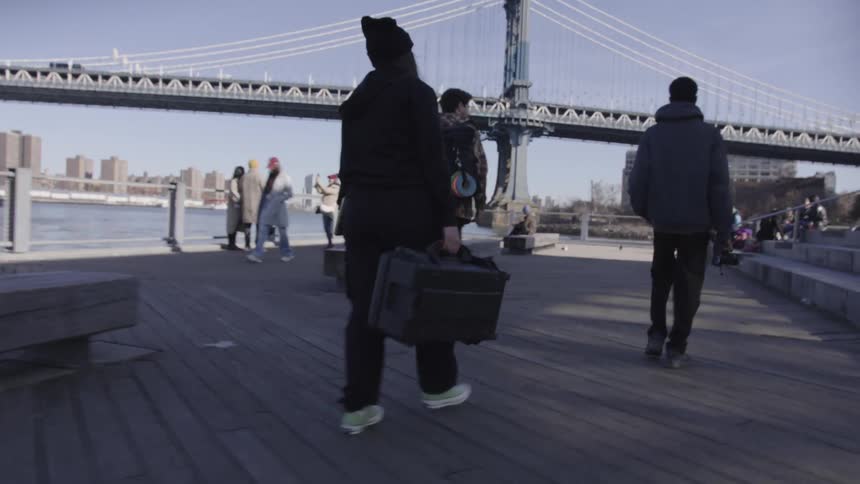 A film crew carrying cameras and equipment in a pier next to a large suspension bridge