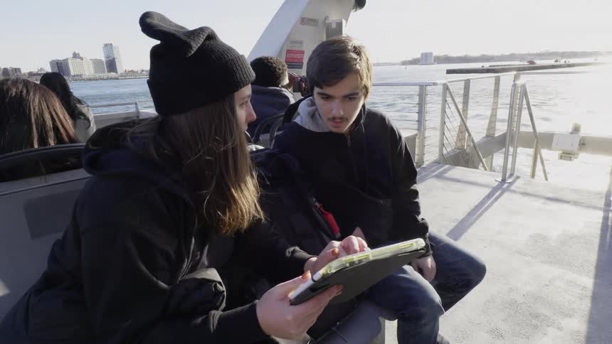Two people seated on the deck of a ferry comparing notes on a tablet