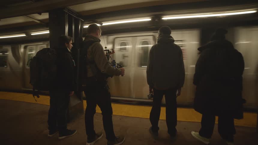 People wait for a subway on the platform one of them is filming the others with a hand held set up