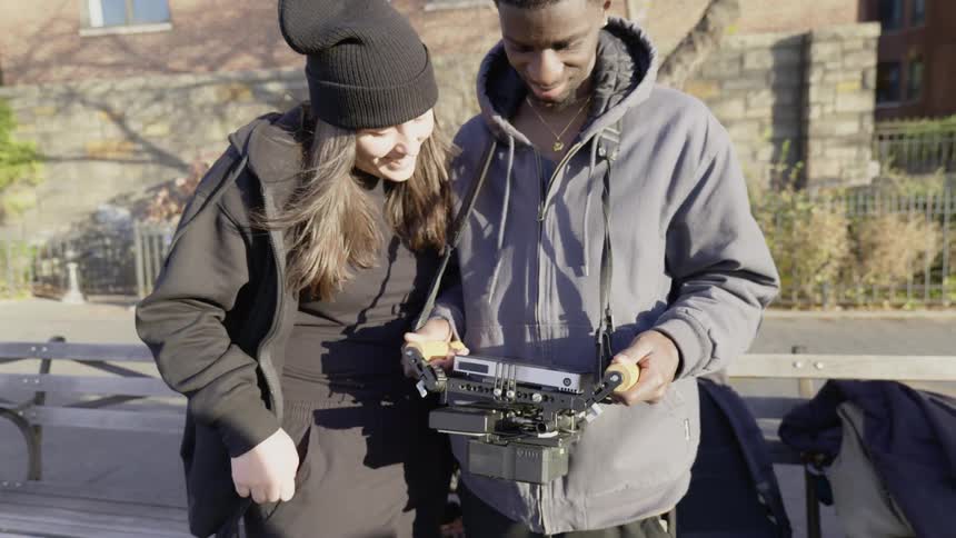 A young woman in a black beanie and hoodie smiling as she points at a device held by a young man in a gray hoodie both engaged in an outdoor setting with warm sunlight