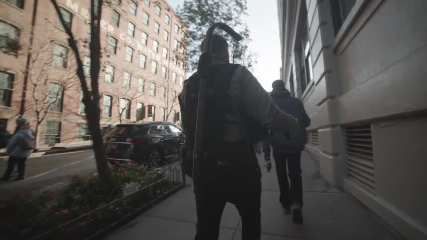 A film crew carrying equipment walks along a city sidewalk at morning