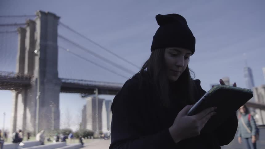 A woman is standing near a bridge in a city typing on a tablet
