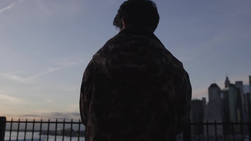 A man wearing a patterned jacket holds a camera standing with their back to the viewer looking out over a city skyline against a twilight sky