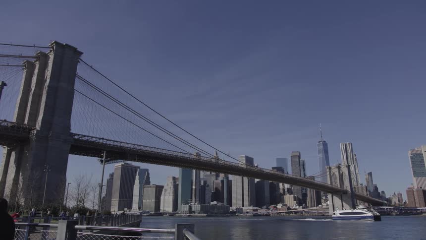 The brooklyn bridge spans across a waterway towards the manhattan skyline under a clear blue sky